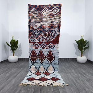 May include: A rectangular, hand-woven rug with a geometric pattern. The rug features a color palette of red, white, blue, and cream. Diamond and square shapes are prominent in the design. The rug is displayed on a gray wooden floor.