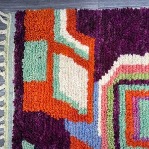 May include: A colorful, geometric rug with a fringe border. The rug features shades of orange, purple, green, white, and blue.