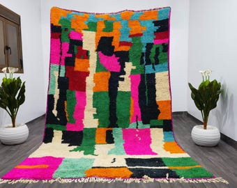 Può includere: Un vivace tappeto fatto a mano con un design patchwork. Il tappeto presenta un'audace tavolozza di colori rosa, arancione, verde, nero e crema. Il tappeto rettangolare è esposto contro un muro bianco, mostrando il suo motivo geometrico.