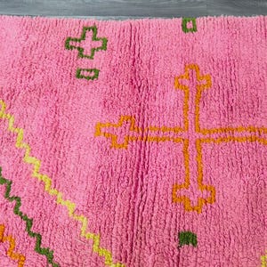 May include: Pink rug with a geometric pattern in orange, green, and yellow. The pattern includes a cross shape in the center.