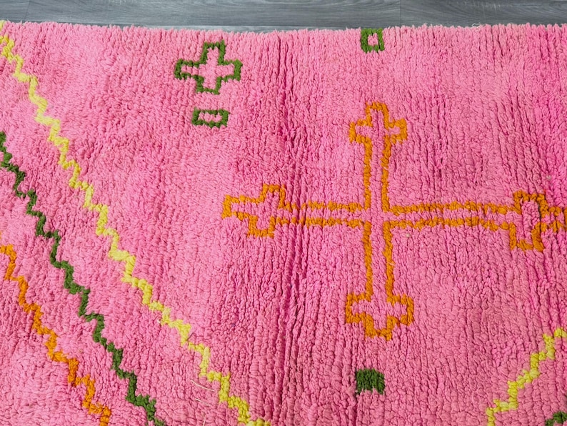 May include: Pink rug with a geometric pattern in orange, green, and yellow. The pattern includes a cross shape in the center.