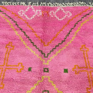 May include: Pink Moroccan rug with a geometric pattern in shades of orange, yellow, and green. The rug has a fluffy texture and a fringe border.