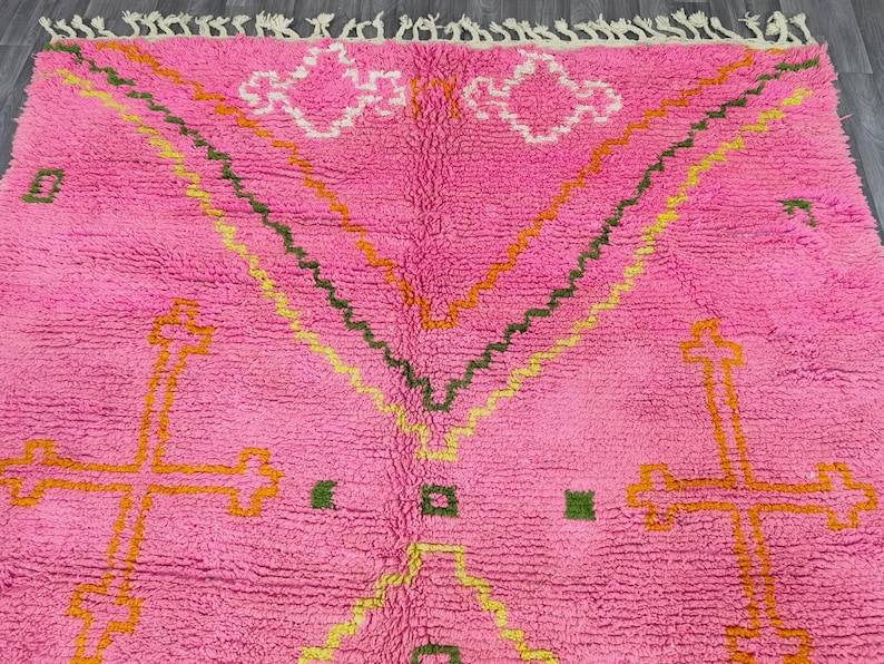 May include: Pink Moroccan rug with a geometric pattern in shades of orange, yellow, and green. The rug has a fluffy texture and a fringe border.