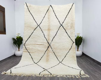 May include: Cream-coloured, rectangular rug featuring a geometric diamond pattern in black. The rug has a shaggy texture and a fringe along the bottom edge. Two potted plants flank the rug, adding greenery to the scene.