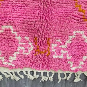May include: Pink and white Moroccan rug with a geometric pattern and orange accents. The rug has a fringe border.