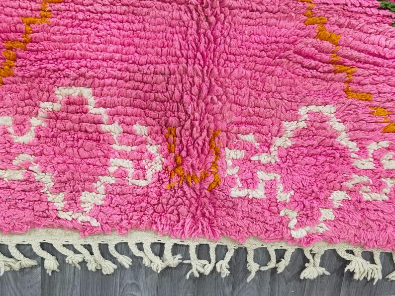 May include: Pink and white Moroccan rug with a geometric pattern and orange accents. The rug has a fringe border.