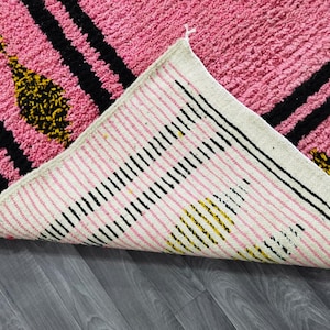 Op de afbeelding: Een roze en wit tapijt met zwarte en gele strepen. Het tapijt heeft een franje rand en is gemaakt van een zacht, pluche materiaal.