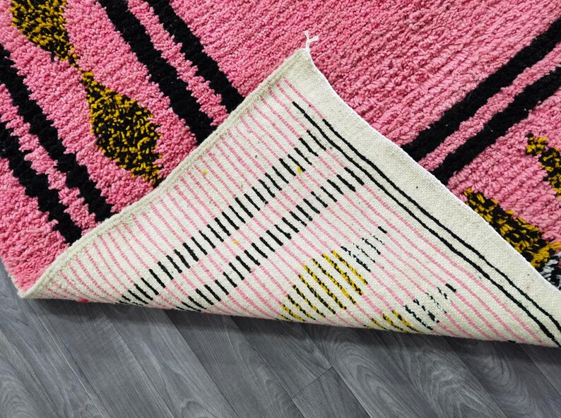 Op de afbeelding: Een roze en wit tapijt met zwarte en gele strepen. Het tapijt heeft een franje rand en is gemaakt van een zacht, pluche materiaal.