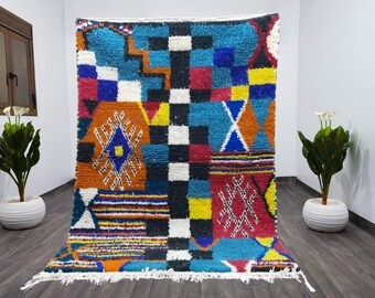 Può includere: Un tappeto marocchino colorato con motivi geometrici in tonalità di blu, rosso, arancio, giallo e bianco. Il tappeto ha un bordo a frange ed è fatto di lana.