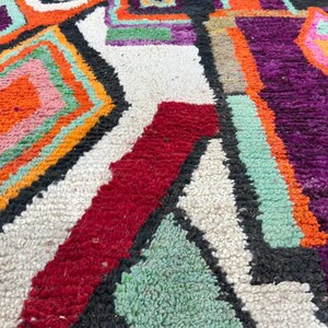 May include: Close-up of a colorful, hand-woven rug with geometric patterns in shades of orange, pink, green, purple, and black. The rug has a white background and a textured, fuzzy surface.