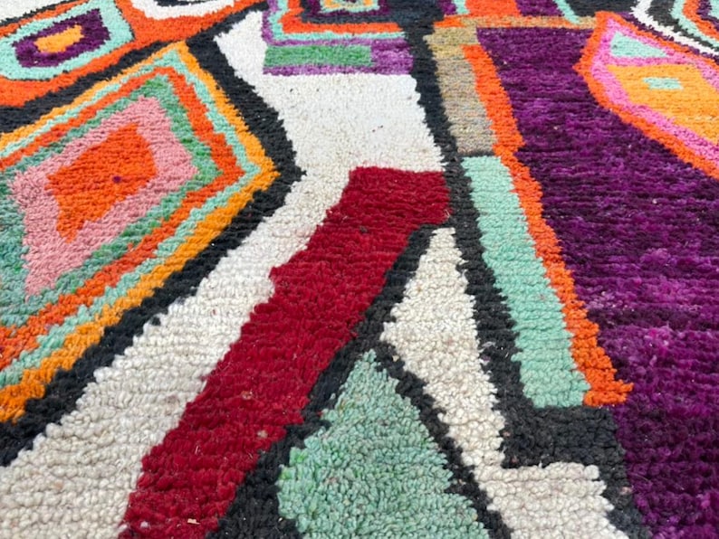 May include: Close-up of a colorful, hand-woven rug with geometric patterns in shades of orange, pink, green, purple, and black. The rug has a white background and a textured, fuzzy surface.