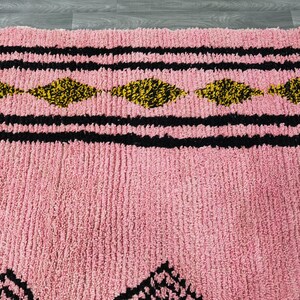 Op de afbeelding: Close-up van een roze en zwart gepatroneerd tapijt met een pluche textuur. Het tapijt heeft een geometrisch ontwerp met zwarte en gele diamanten.