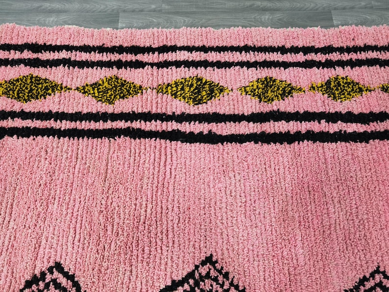 Op de afbeelding: Close-up van een roze en zwart gepatroneerd tapijt met een pluche textuur. Het tapijt heeft een geometrisch ontwerp met zwarte en gele diamanten.