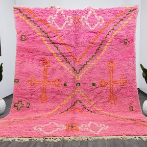 May include: A pink Moroccan rug with a geometric pattern featuring crosses and diamond shapes. The rug has a fringe border and is made of wool.
