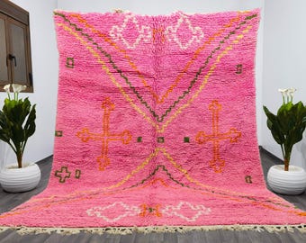 May include: A pink Moroccan rug with a geometric pattern featuring crosses and diamond shapes. The rug has a fringe border and is made of wool.