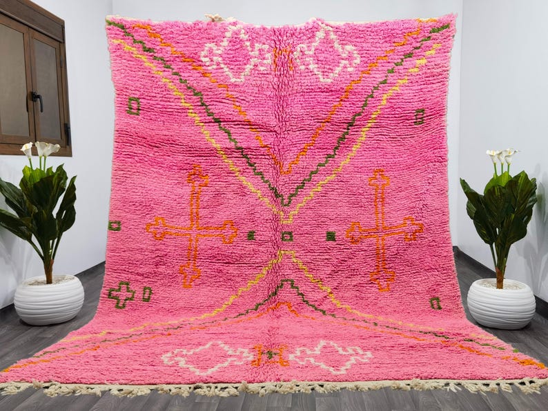 May include: A pink Moroccan rug with a geometric pattern featuring crosses and diamond shapes. The rug has a fringe border and is made of wool.