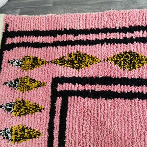 Op de afbeelding: Roze kleed met een zwarte en witte rand en gele en zwarte geometrische ontwerpen.