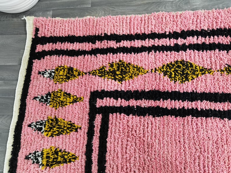 Op de afbeelding: Roze kleed met een zwarte en witte rand en gele en zwarte geometrische ontwerpen.