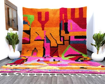 Op de afbeelding: Een kleurrijk abstract tapijt met geometrische vormen in tinten oranje, roze, groen en blauw. Het tapijt heeft een franje rand en is gemaakt van wol.
