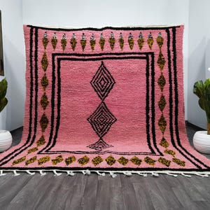 Op de afbeelding: Een roze Marokkaanse tapijt met een zwart en gouden ruitpatroon. Het tapijt heeft een franje rand.