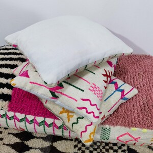 Puede incluir: Una pila de cojines decorativos en varios estilos y colores. El coj&iacute;n superior es blanco, mientras que otros presentan patrones geom&eacute;tricos y texturas en rosa, negro y crema. Los cojines est&aacute;n colocados sobre una alfombra estampada.