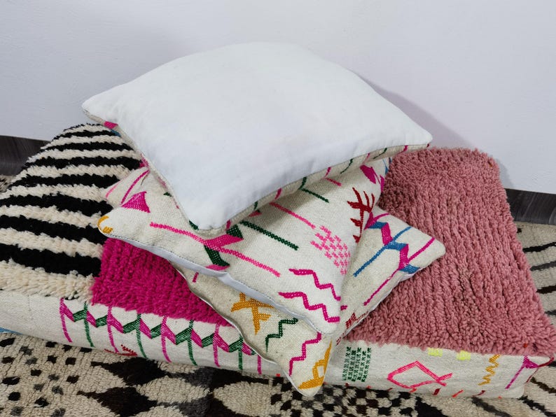 Puede incluir: Una pila de cojines decorativos en varios estilos y colores. El coj&iacute;n superior es blanco, mientras que otros presentan patrones geom&eacute;tricos y texturas en rosa, negro y crema. Los cojines est&aacute;n colocados sobre una alfombra estampada.