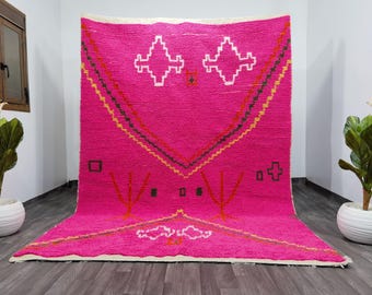May include: A vibrant pink rug with geometric patterns. The rug features white, orange, yellow, and black designs. The central design includes two white diamond shapes. The rug is made of a thick, textured material.