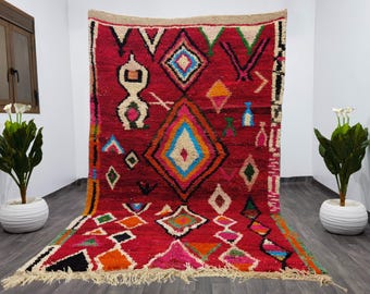 May include: A vibrant, hand-woven rug with a red background and geometric patterns in various colors. The design includes diamonds, triangles, and other shapes in shades of blue, orange, green, and white. The rug has a fringed edge.
