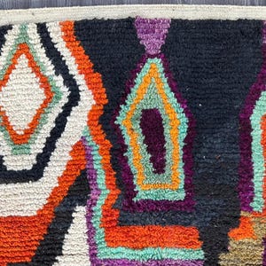 May include: Close-up of a colorful woven rug with geometric patterns in shades of orange, white, black, purple, and green.
