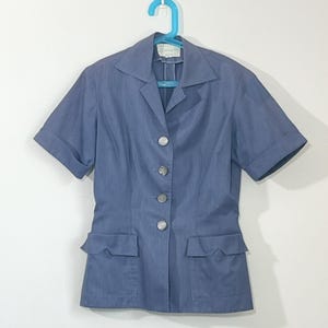 May include: A short-sleeved, light blue blazer with a notched collar and four visible buttons. The blazer has two front pockets and is hanging on a blue hanger. The fabric appears to be a lightweight cotton blend.
