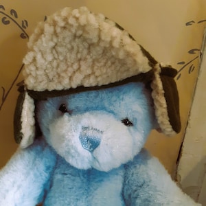 Pode incluir: Um urso de peluche azul claro usando um chapéu de aviador marrom e verde com abas para as orelhas.