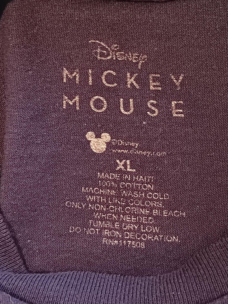 May include: Close-up of a dark blue t-shirt label with the Disney Mickey Mouse logo and text. The label includes washing instructions, size XL, and the text "Made in Haiti, 100% Cotton."