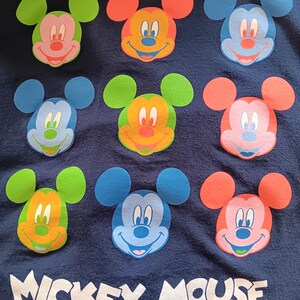 May include: Navy blue fabric featuring nine colorful Mickey Mouse face appliqu&eacute;s. Each Mickey face has a different color scheme, including green, orange, blue, and pink. The words "Mickey Mouse" are printed in white at the bottom.