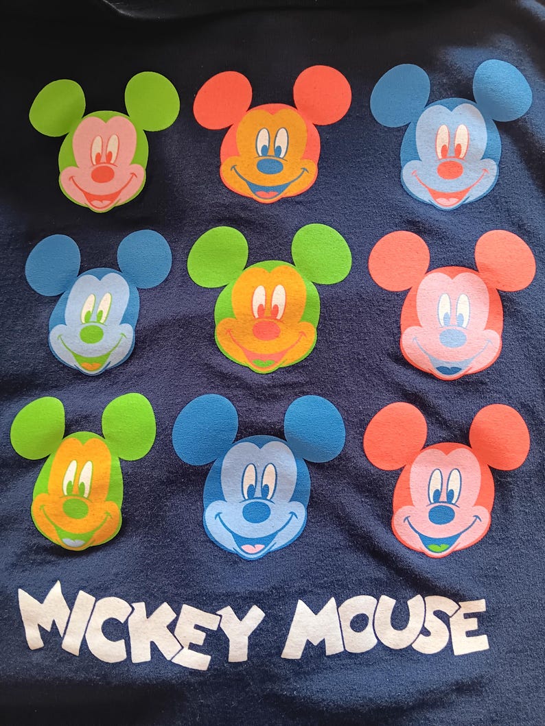 May include: Navy blue fabric featuring nine colorful Mickey Mouse face appliqu&eacute;s. Each Mickey face has a different color scheme, including green, orange, blue, and pink. The words "Mickey Mouse" are printed in white at the bottom.