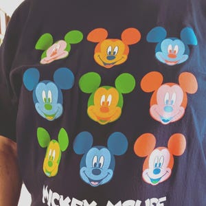 May include: Navy blue t-shirt featuring a colorful Mickey Mouse design. The graphic includes multiple Mickey Mouse faces in various colors, such as orange, blue, and green. The words "Mickey Mouse" are printed in white at the bottom of the shirt.