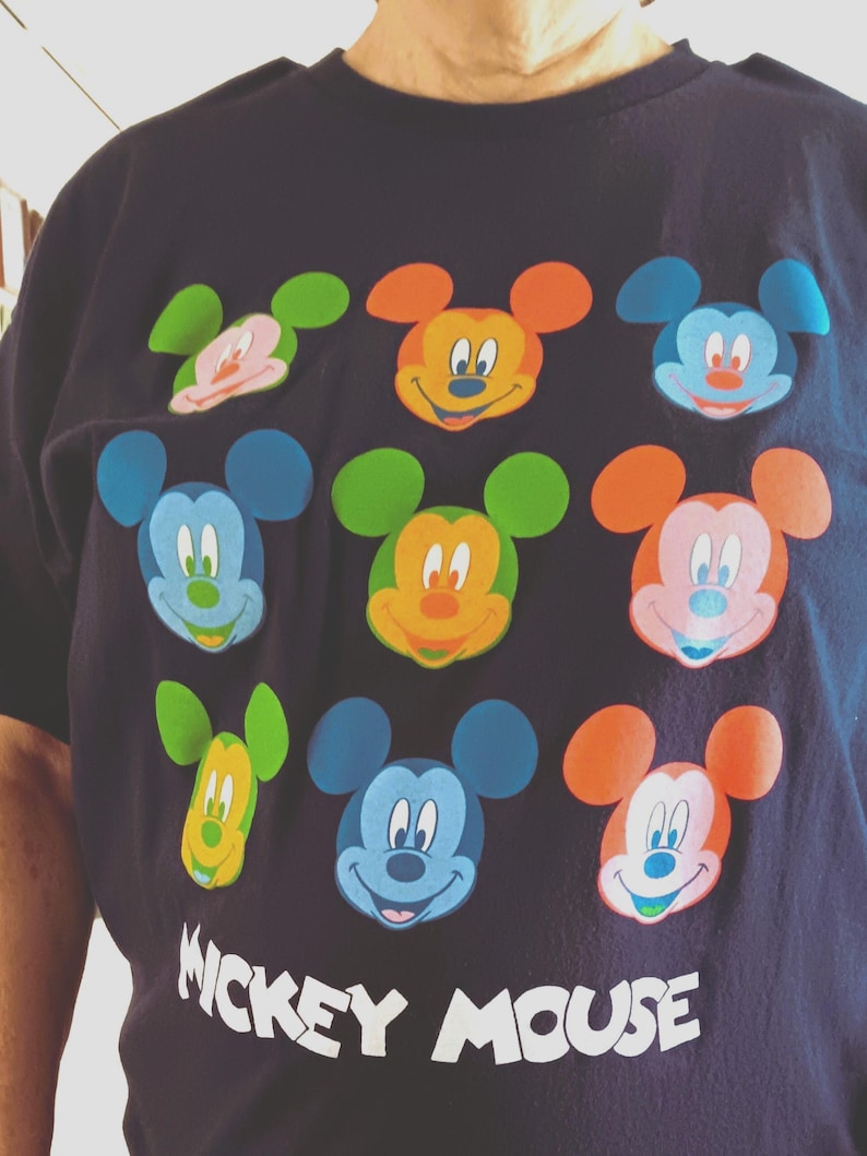 May include: Navy blue t-shirt featuring a colorful Mickey Mouse design. The graphic includes multiple Mickey Mouse faces in various colors, such as orange, blue, and green. The words "Mickey Mouse" are printed in white at the bottom of the shirt.