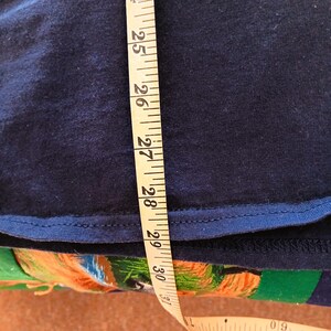 May include: A navy blue garment is measured with a yellow measuring tape. The tape shows measurements from 23 to 31 inches. The garment's edge is visible, with a contrasting blue trim. The background is a colorful fabric.