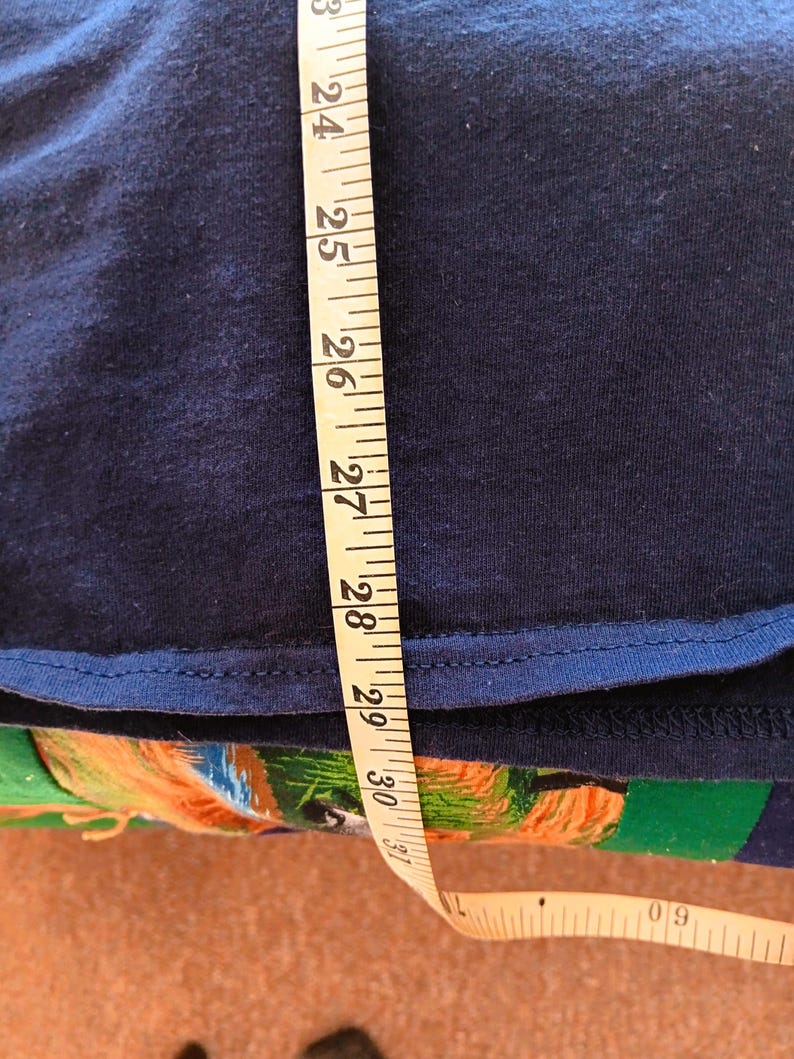 May include: A navy blue garment is measured with a yellow measuring tape. The tape shows measurements from 23 to 31 inches. The garment's edge is visible, with a contrasting blue trim. The background is a colorful fabric.