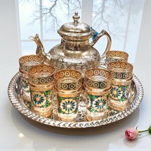 Könnte beinhalten: Ein silbernes Teeservice mit einer Teekanne und mehreren dekorativen Gläsern, alles auf einem silbernen Tablett angeordnet. Die Gläser haben ein gold-blaues Blumenmuster. Die Teekanne ist aufwendig detailliert.