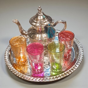 Puede incluir: Un juego de té plateado con una tetera y cinco vasos coloridos en una bandeja a juego. Los vasos son de color naranja, rosa, verde y rojo. La tetera tiene un diseño decorativo y una borla.