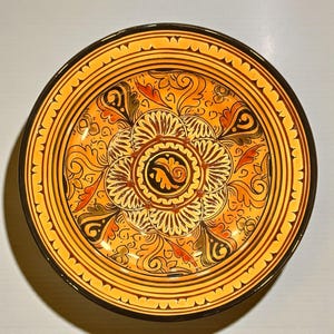 Puede incluir: Un plato redondo de cerámica decorativo con un diseño floral vibrante. El plato presenta un fondo naranja cálido con intrincados patrones en negro, marrón y rojo. El borde es de color marrón oscuro y el plato tiene un borde festoneado.