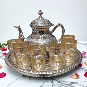 Puede incluir: Un juego de té plateado con una tetera y seis vasos, todo sobre una bandeja plateada. Los vasos tienen un patrón geométrico dorado. La tetera tiene detalles intrincados y una tapa decorativa. El juego está dispuesto sobre una superficie blanca con pétalos de rosa esparcidos.