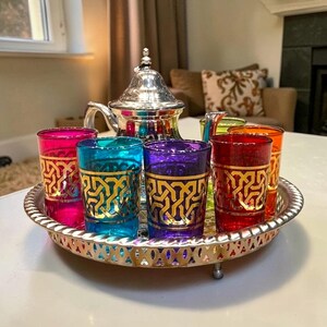 Peut inclure: Une théière en métal argenté avec un couvercle pointu est posée sur un plateau rond assorti, accompagnée de six petits verres aux couleurs vives comme le magenta, le turquoise, le violet, le rouge, l'orange et le vert, ornés de motifs dorés.