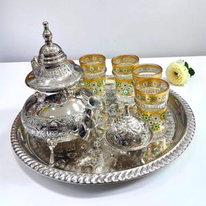 May include: A silver-toned metal tea set featuring a large teapot with a pointed lid, a smaller lidded sugar bowl, and four glass cups with gold rims and colorful floral patterns, all arranged on a large, round, embossed silver-toned metal tray.