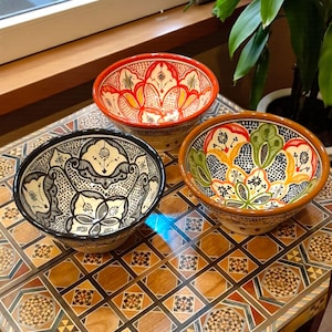 Peut inclure: Trois bols en céramique décoratifs avec des motifs floraux complexes. Les bols présentent une variété de couleurs, dont le rouge, le noir et le vert, avec des motifs détaillés. Les bols sont exposés sur une surface en bois avec un motif de mosaïque.
