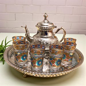 Könnte beinhalten: Ein silbernes Teeservice mit einer Teekanne und sechs Gläsern, alles auf einem silbernen Tablett. Die Gläser haben bunte Blumenmuster in Blau, Rot und Gold. Die Teekanne hat aufwendige Designs.