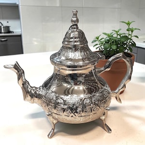 May include: Ornate silver teapot with intricate embossed patterns, a curved spout, and a decorative handle. The teapot sits on three small feet and has a domed lid with a finial. A small potted plant is in the background.