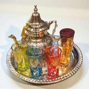 Può includere: Un set da tè argentato con una teiera decorativa e sei bicchieri colorati su un vassoio d'argento. I bicchieri sono gialli, verdi, blu, rossi e arancioni. La teiera ha un nappa e dettagli ornati.