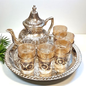 Può includere: Un servizio da tè argentato con una teiera decorativa, sei bicchieri e un vassoio rotondo. I bicchieri presentano un motivo floreale oro e nero. La teiera e il vassoio hanno intricati motivi in rilievo. Il set è progettato per servire il tè.