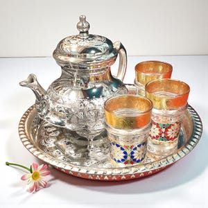 Könnte beinhalten: Ein silbernes Teeservice mit einer dekorativen Teekanne, drei Gläsern und einem Serviertablett. Die Gläser haben goldene Ränder und bunte Blumenmuster. Das Tablett hat einen roten Rand und eine Gänseblümchenblüte an der Seite.